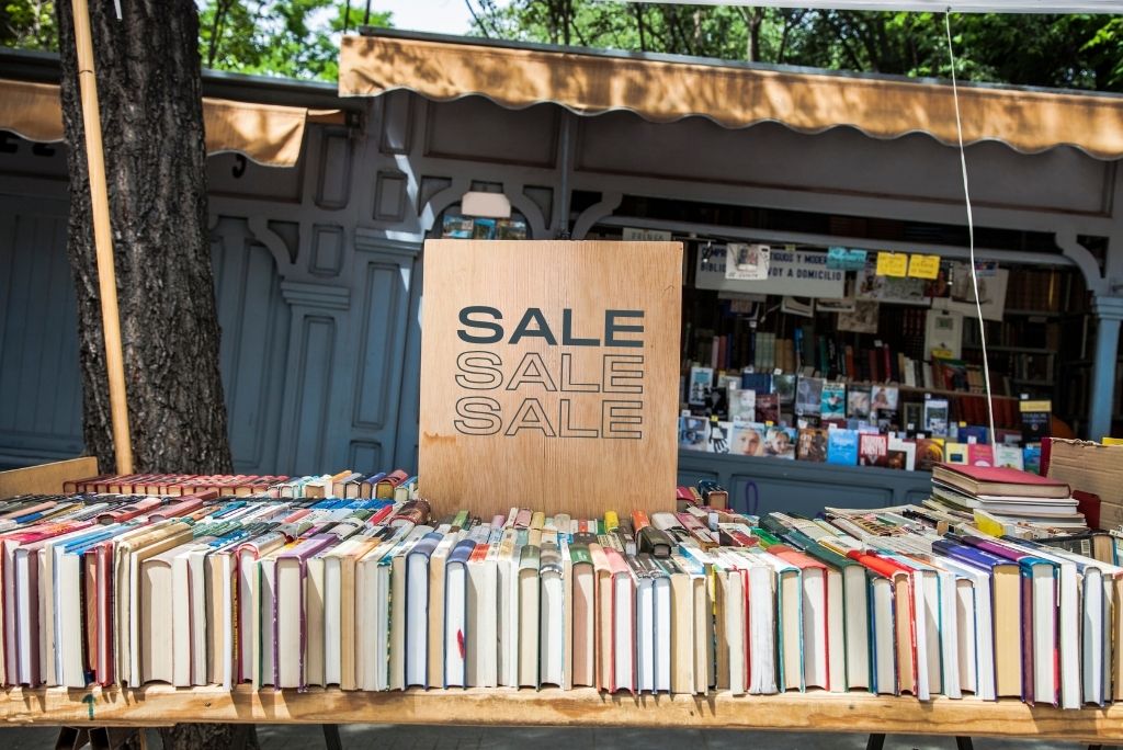 Outdoor book sale with rows of books on a table and a large wooden 'SALE' sign in front, under a canopy of trees.