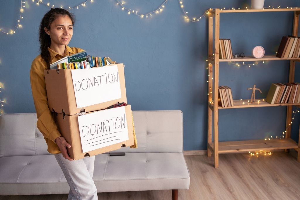 A woman smiling, holding two boxes labeled "DONATION" full of books, with a bookshelf in the background.