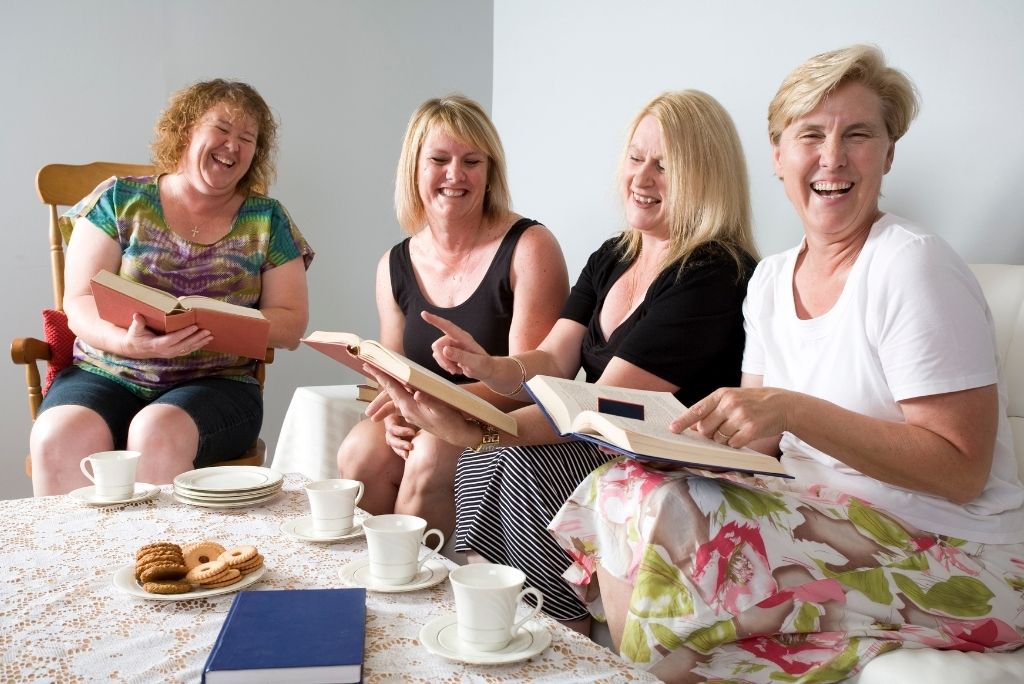 Group of cheerful women enjoying a book club discussion with tea and biscuits.