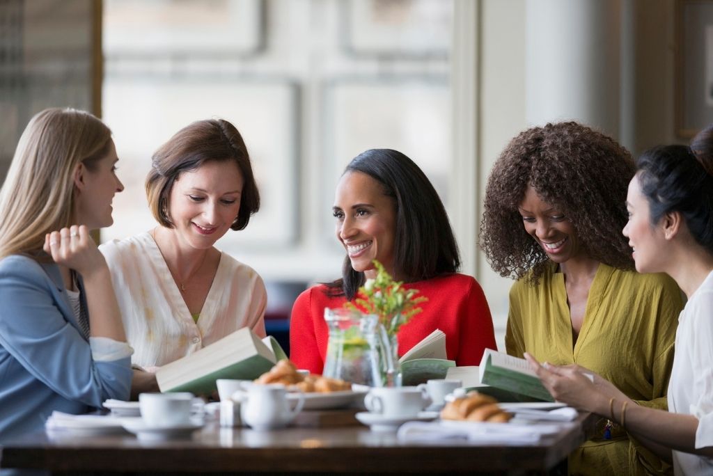 Diverse group of women engaged in a lively book club discussion at a cafe table.