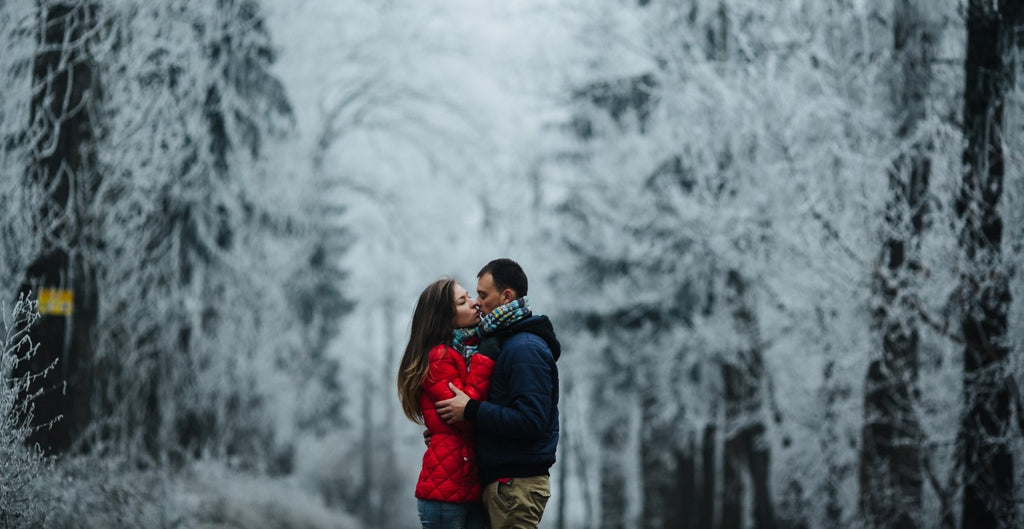 A couple shares a kiss on a snow-lined path in a wintery forest.