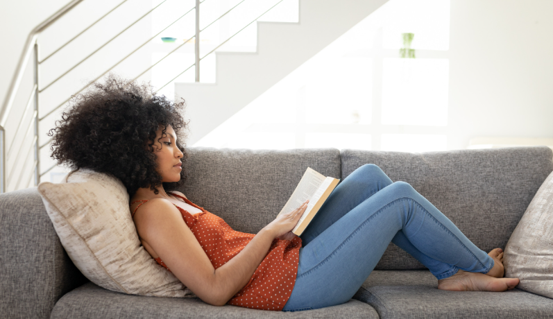 A black girl with curly hair is lying on a sofa and reading a book in her lap.