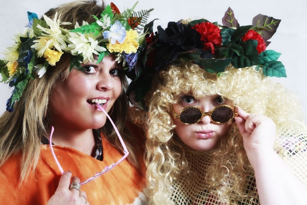 Two women in floral wreaths and quirky costumes, evoking a fun and playful atmosphere.