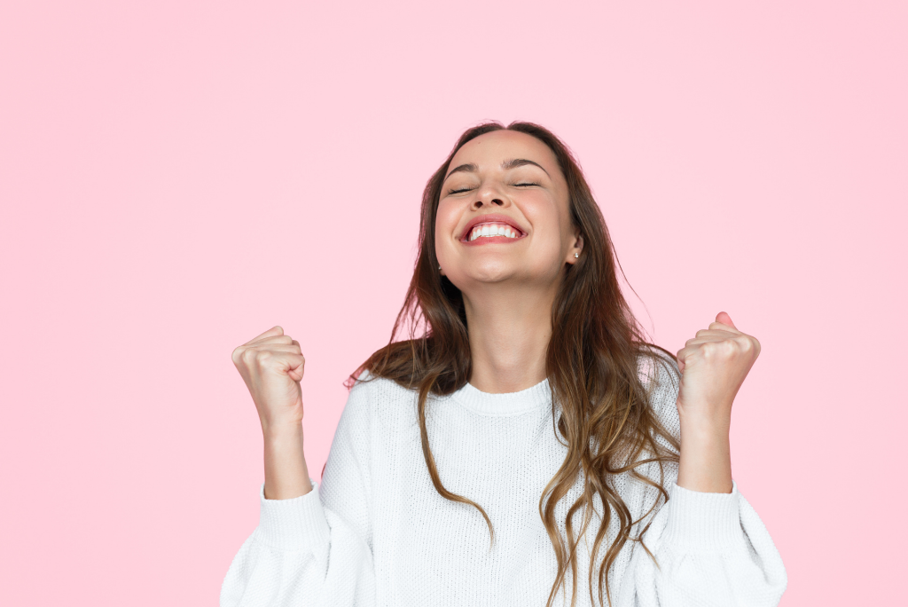 Overjoyed woman with eyes closed and fists pumped in celebration against pink backdrop.
