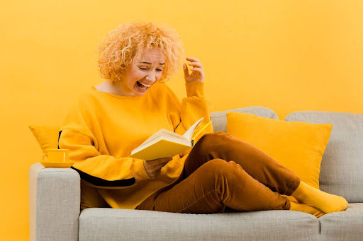 Alt text: A woman with curly blonde hair, wearing a vibrant yellow sweater, laughs while reading a book, comfortably seated on a gray couch with matching yellow cushions against a yellow background.