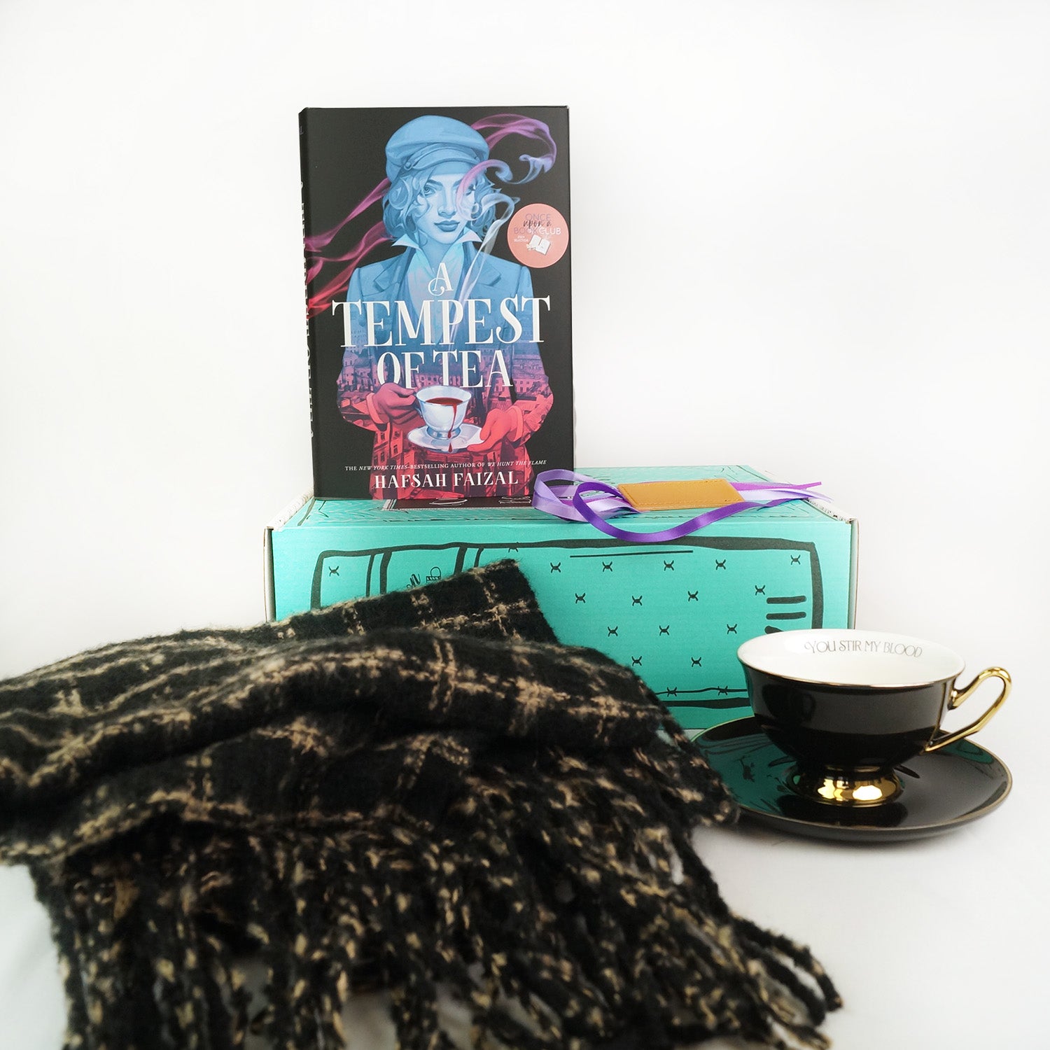 A hardcover edition of A Tempest of Tea is next to a wrapped gift labeled with a page number sitting on top of a green Once Upon a Book Club box. In front are a black and gold oversized scarf and black teacup plus plate.