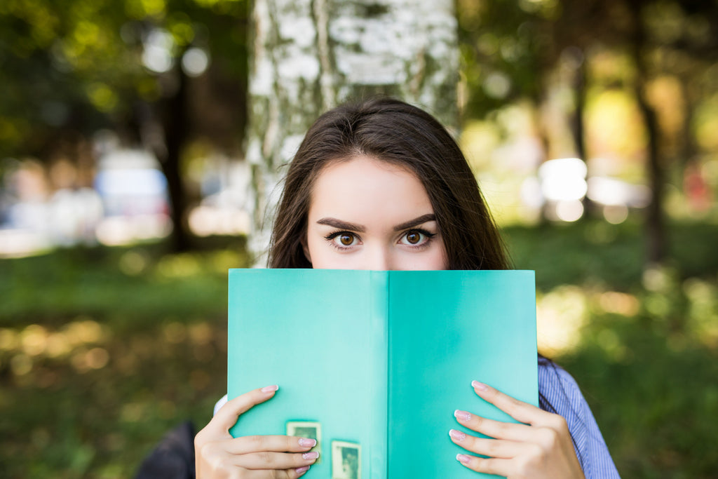 A woman with striking eyes peeks over the top of a teal book, half her face hidden, suggesting curiosity and intrigue, set against a blurred natural park background.