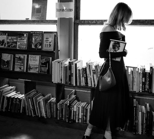 A woman in a black dress stands reading a book in a library with shelves filled with a variety of books, captured in a candid, serene black and white photo.