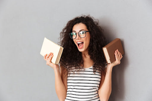 An exuberant woman with curly hair and glasses, wearing a striped shirt, excitedly holds a book in each hand against a gray background. Her wide-eyed expression and open-mouthed smile convey a sense of joyful surprise or enthusiasm.