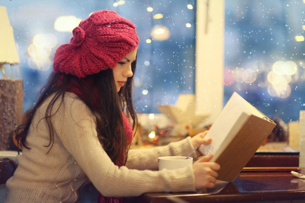 Woman in a cozy hat engrossed in a book with a cup of tea, snowflakes outside.