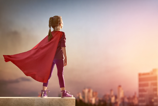 A child in a red cape stands heroically on a ledge overlooking the city at sunset, evoking empowerment.