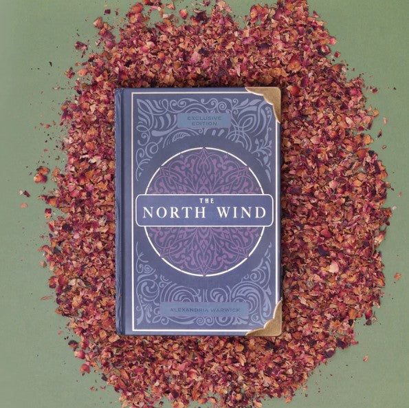 The book 'The North Wind,' written by Alexandria Warwick, is placed atop a bed of dried red rose petals.