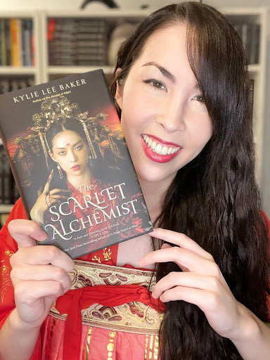 A cheerful woman with long hair holds up a copy of 'The Scarlet Alchemist' by Kylie Lee Baker, matching the book's cover with her red outfit.