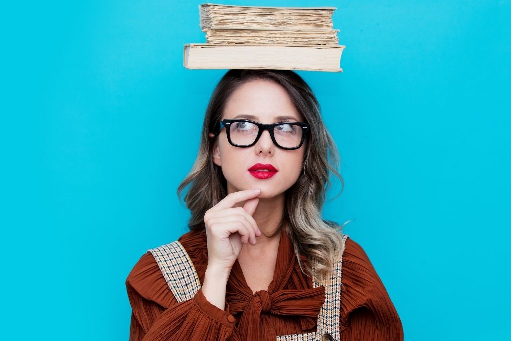 Thoughtful woman with glasses balancing a stack of books on her head against a blue background.