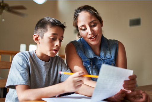 A boy focuses on writing as a woman, likely his mother, guides him, suggesting a creative writing session at home.