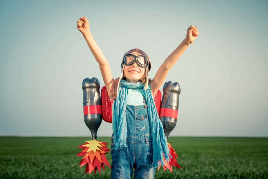 A child in a jetpack costume with arms raised high in a field.