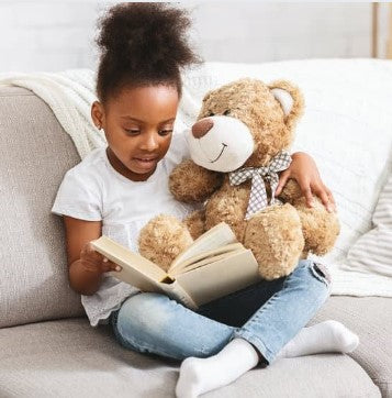 A black baby girl is sitting on a white sofa, holding a teddy bear in her hands and reading a book in her lap.