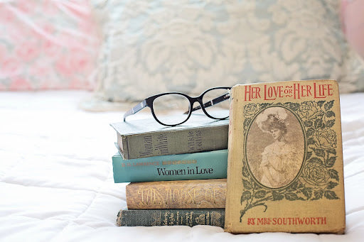 A stack of classic novels including "Women in Love" and "The House of Life," topped with a pair of black-rimmed glasses, set against a cozy bedroom background with decorative pillows.