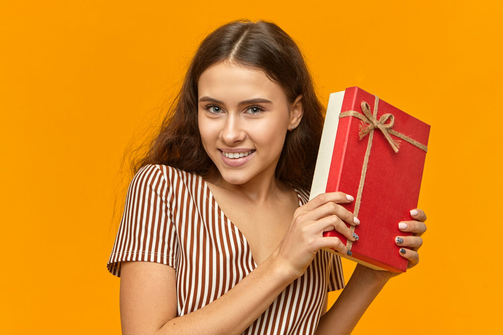 Smiling woman holding a red-wrapped book with a bow on a yellow background.