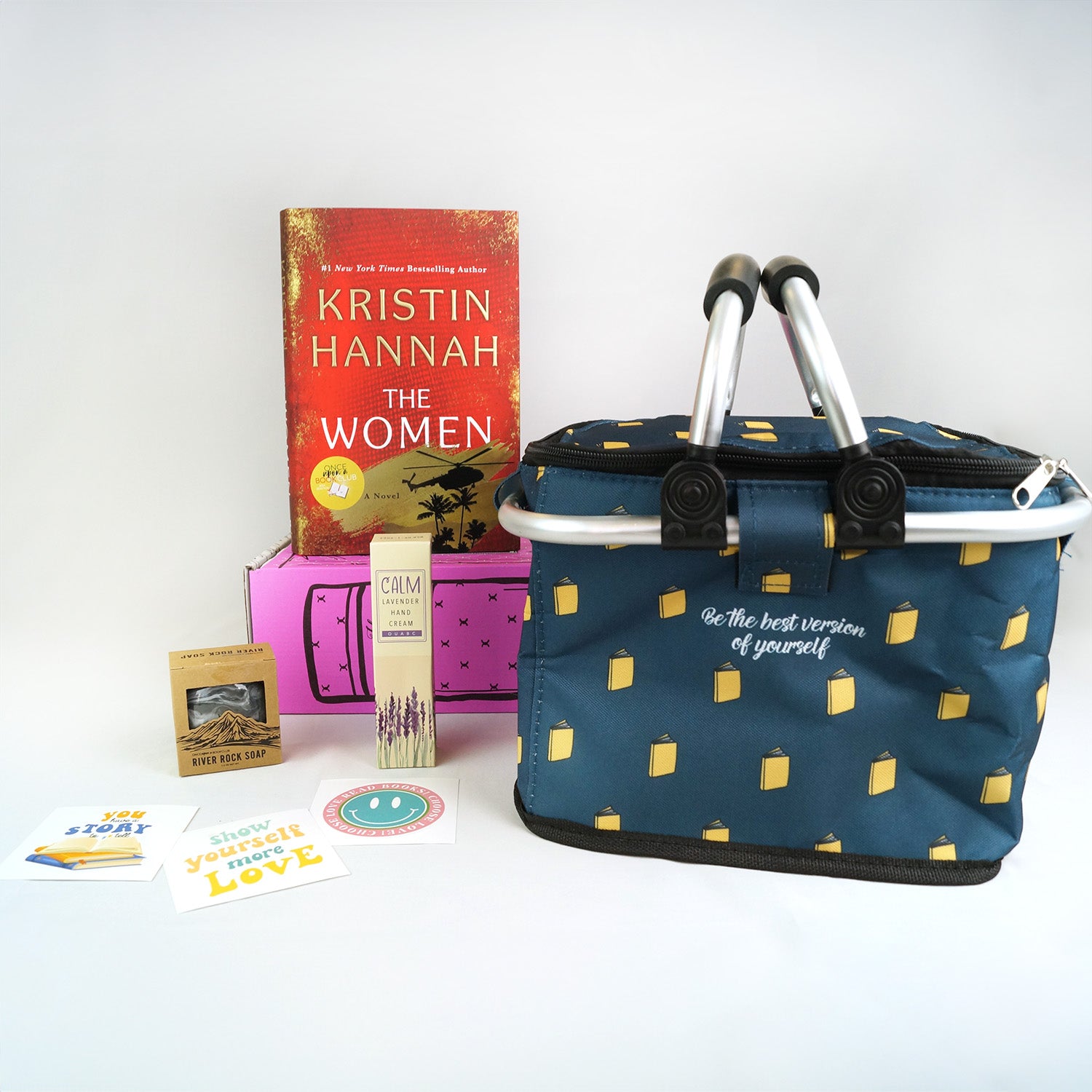 A hardcover edition of The Women stands on a pink Once Upon a Book Club box. In front are a brown square box, three stickers, a box with lavender on it, and a navy blue cooler with yellow books on it and white lettering that says Be the Best Version of Yourself