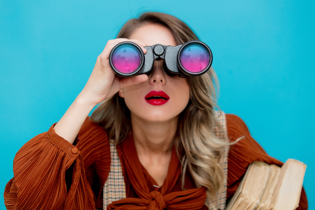 Woman looking through binoculars with a book, against a blue background.