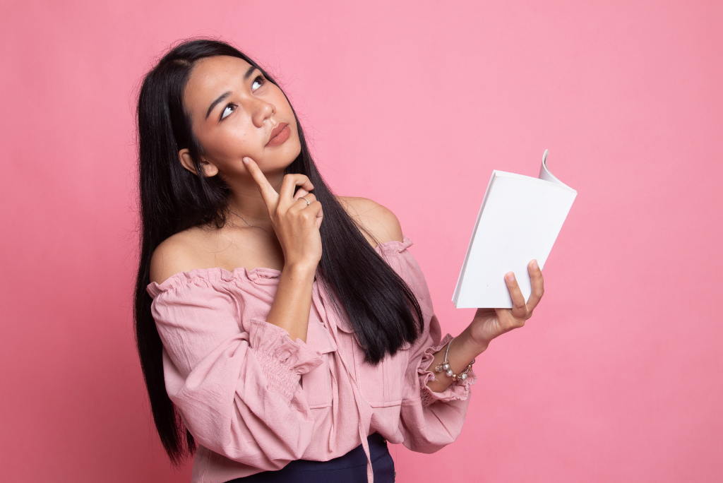 A thoughtful woman holding a blank book with a pink background.