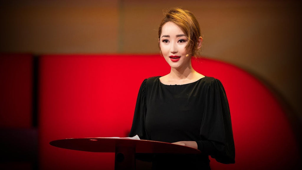 A woman speaking confidently at a podium, wearing a black dress with a microphone headset, set against a bright red background.