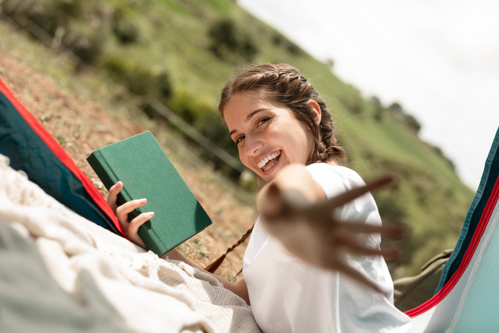 Smiling woman with book reaching out from hammock in nature.