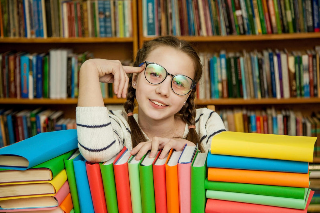 A young girl with glasses smiling behind a stack of colorful books.
