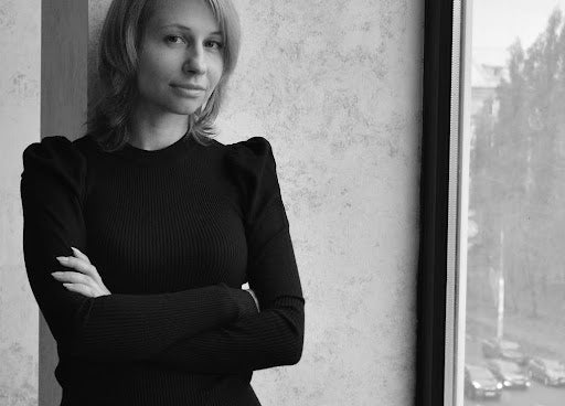 A black-and-white portrait of a confident woman with crossed arms, wearing a black turtleneck, standing by a window with a contemplative expression, overlooking a blurred cityscape.