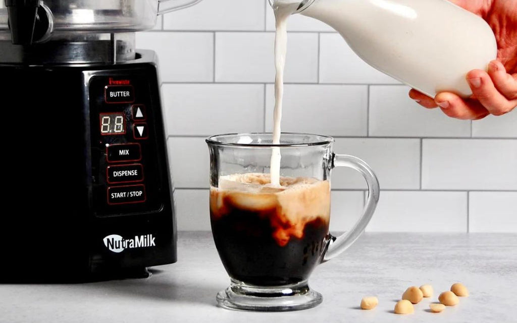 Macadamia milk is being poured into a glass of cold brew coffee sitting next to a NutraMilk processor.