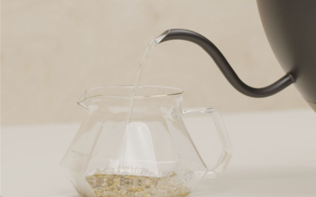 Hot water is slowly pouring out of a black artisan gooseneck kettle into a glass server filled with loose leaf tea.