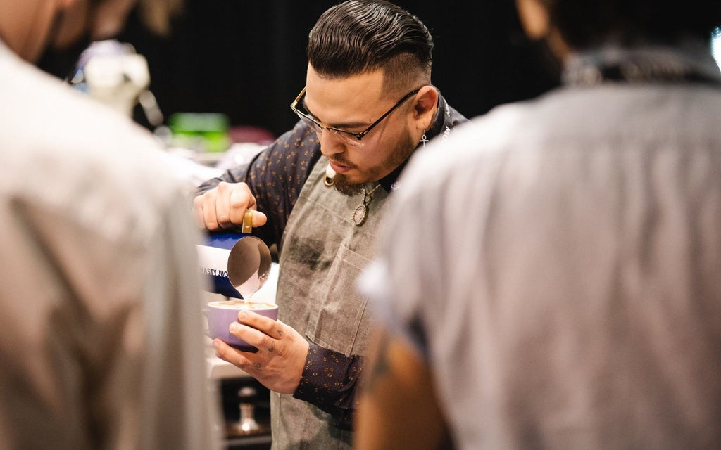 Proph is pouring latte art from a Nasty Jug while competition judges watch on.