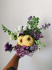 A bouquet of flowers - white, yellow, green, purple - held up against a beige wall