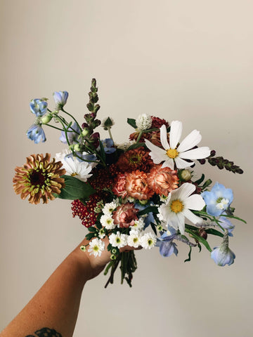 A bouquet of flowers - with blues, whites, and peachy tones held up against a beige wall