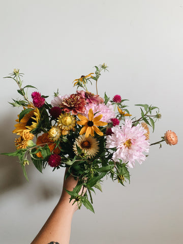 A bouquet of flowers - yellow, light pink, orange - held up against a beige wall