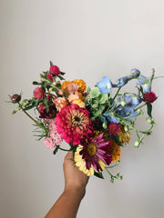 A bouquet of flowers - pink, blue, yellow, green, purple - held up against a beige wall
