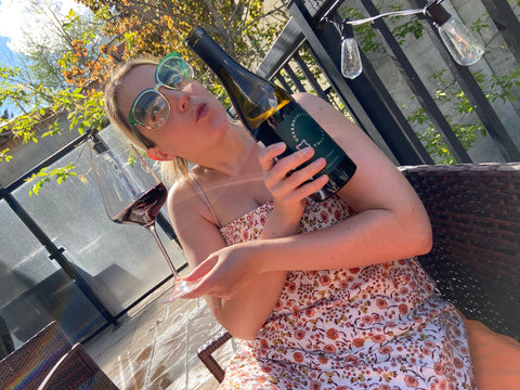 Jen holding a bottle of wine and a glass sitting outside in the sun