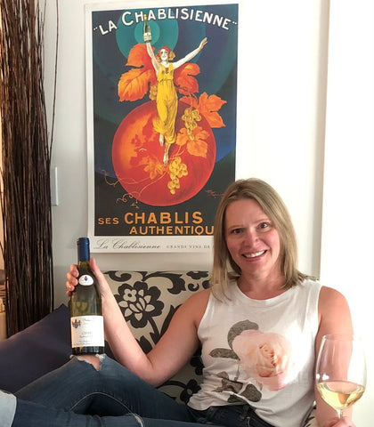 Nathalie holding a bottle of wine and glass sitting in front of a Chablis poster
