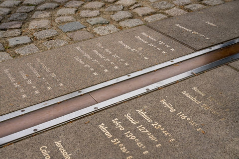 The Greenwich Mean Time, prime meridian in England. There is a brown line bordered by metal strips with cities and their times running from the bottom left to the top right.