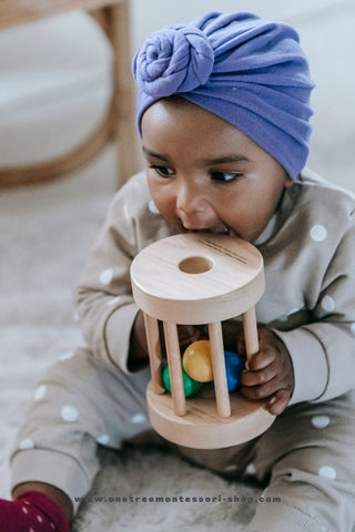 A Black baby with a blue turban is biting a wooden rattle toy.