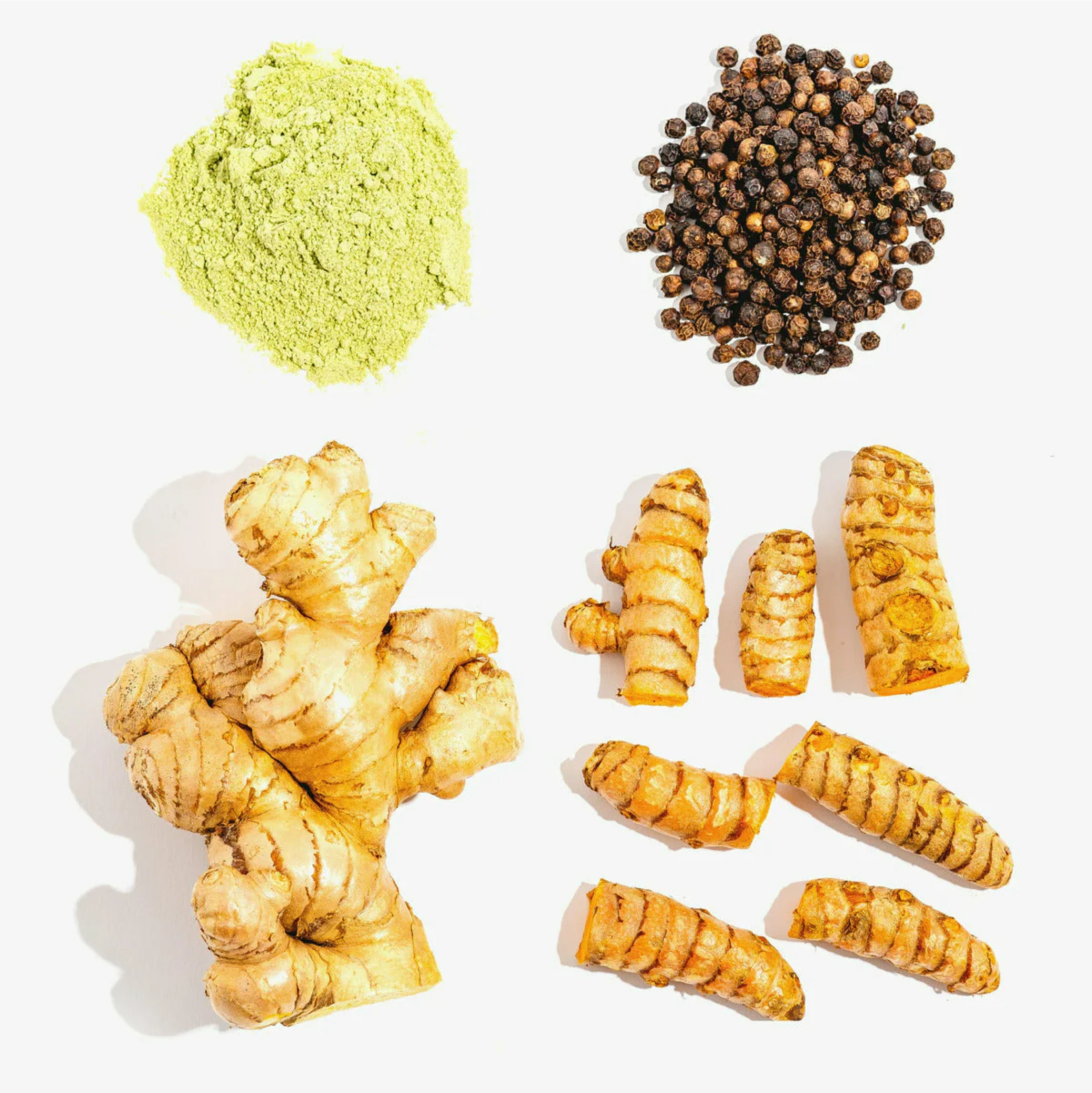 Assorted spices: green powder, black peppercorns, ginger root, and turmeric root against a white background.