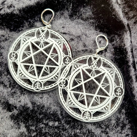 A pair of laser cut clear acrylic earrings in the shape of two circles etched with white symbols from a summoning circle. With silver plated latch hooks. Seen on a grey velvet background 
