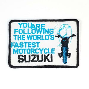 4" x 3" 1970s vintage deadstock (NOS) Suzuki patch in off-white twill with turquoise blue and black embroidery depicting a head-on view of a bike and "YOU ARE FOLLOWING THE WORLD'S FASTEST MOTORCYCLE" text