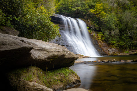     Silver Run Falls Trailhead     There are many pull-outs along this highway that lead to dozens of little known trails in the Nantahala National Forest which end with magnificent mountain vistas or at jaw dropping waterfalls. If you see a random car parked on the side of the road here chances are they’re chasing waterfalls. For this particular hike, we drove 4 miles south of Cashiers until spotting the bright yellow Forest Service sign for Silver Run Falls. Google Maps has the trailhead in their directory so getting here was a breeze. Being early in the evening, we had the place all to ourselves which made it that much more special. At only 0.16 miles long, this trail has one of the best bangs for your buck for all of the scenery that gets packed into such a short duration.      Right off the bat, the gravel trail enters the forest and cuts through an understory of rhododendrons and pines filled with blooming blueberries, mountain laurel, and all other kinds of wildflowers you might expect in early May. The musky scent of wet pine needles mixed with the sweet perfume of the laurels is what I associate most with hiking in the Appalachian Mountains. You’ll pass a small side trail to the left that leads off-trail to another waterfall most visitors are unaware exists, Upper Silver Run Falls. This 20 ft waterfall upstream from the main Silver Run Falls is beautiful in its own right, but requires a hefty hike and dicey vertical scramble to reach. HikingWNC has a great article on this waterfall and how to reach it for anyone interested.     Crossing the Whitewater River     A few short minutes in and you’ll reach the walking bridge spanning the Whitewater River. Up until recently this crossing was merely a simple log, but for safety reasons the Forest Service constructed this sturdy bridge in its place. At this point upstream the Whitewater River is only a dozen feet wide, hinting little as to how enormous this river becomes by the time it reaches Whitewater Falls. Through the trees one can make out Silver Run Falls and hear its booming sound just a skip and a hop away. After crossing the bridge, the trail abruptly ends at a large rock outcropping on the shores of Silver Run Falls large wading pool.      Silver Run Falls      It may not be the tallest or largest waterfall in the Blue Ridge Mountains, but it sure is beautiful. The picturesque scene here screams Western North Carolina. Most people hangout on this outcropping and its the most photographed view of Silver Run Falls I’ve come across. Following the base of the cliff you can walk right up to the precipice of Silver Run Falls and feel the fine misty spray of its 25 ft drop.  Using some of the larger rocks, I made my way across the stream that drains Silver Run Creek into the Whitewater River and landed on the shore opposite the falls. In just a few short weeks this beach will be packed with swimmers bathing in the cool mountain stream and it’ll be impossible to find parking here.   Silver run falls Nantahala National Forest North Carolina hiking trail waterfalls