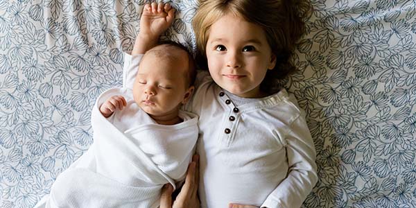 Baby swaddled on bed next to sibling