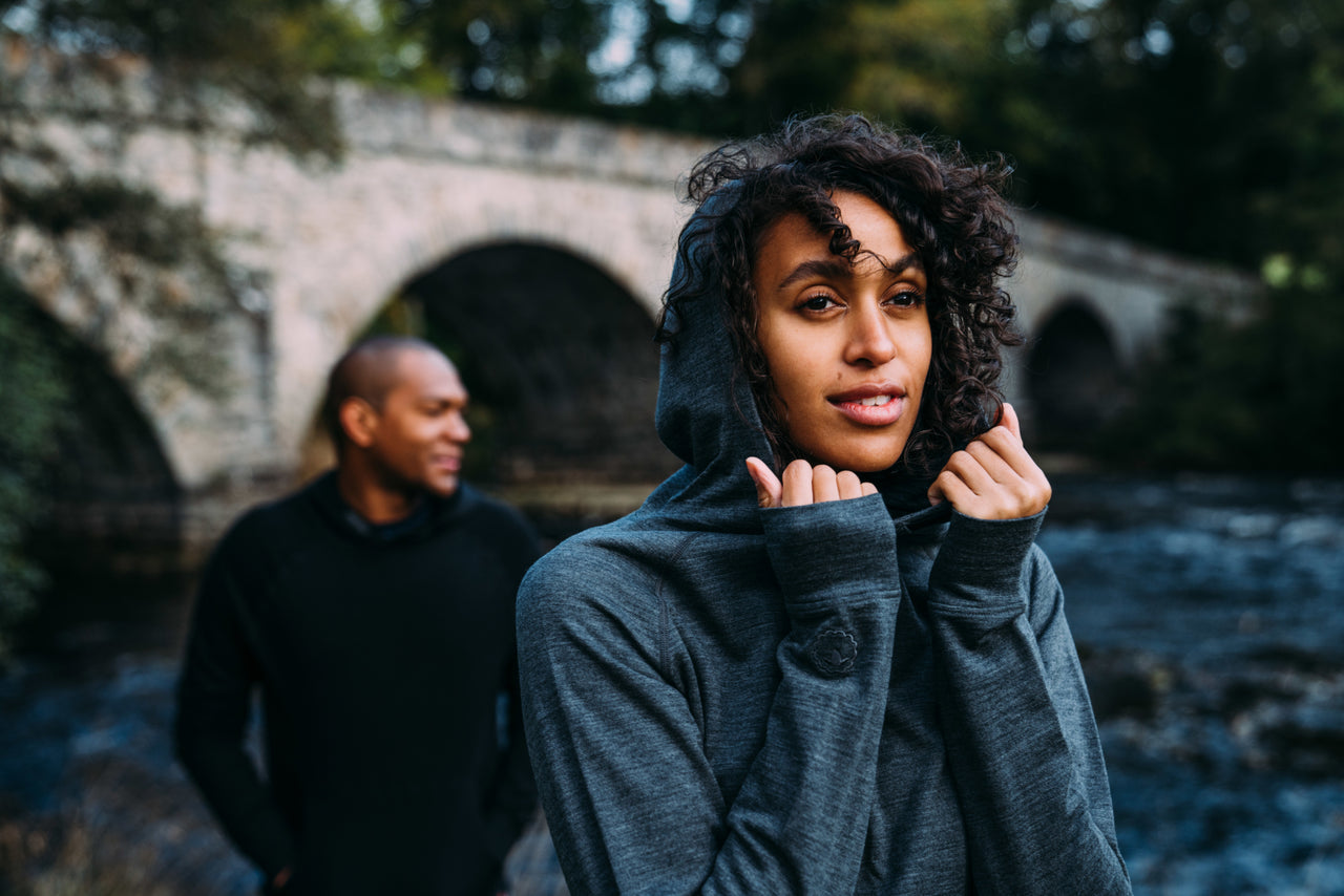 A woman in the foreground looks thoughtfully at the camera, with a historic stone bridge and a flowing river in the background. A man, slightly out of focus, is standing behind her looking away from the camera. Both are surrounded by a tranquil woodland scene and are wearing Isobaa 260 Casual Hoodies.