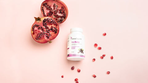 Eine Flasche SanaExpert Acai Berry Nahrungsergänzungsmittel liegt auf einer weichen rosa Oberfläche, umgeben von Granatapfelkernen und einer halben Frucht.