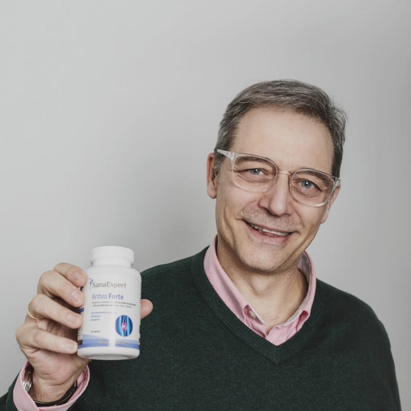 Ein lächelnder Mann mittleren Alters in einem grünen Pullover präsentiert stolz eine Flasche SanaExpert Arthro Forte, betonend auf persönliches Wohlbefinden und Vertrauen in das Produkt.