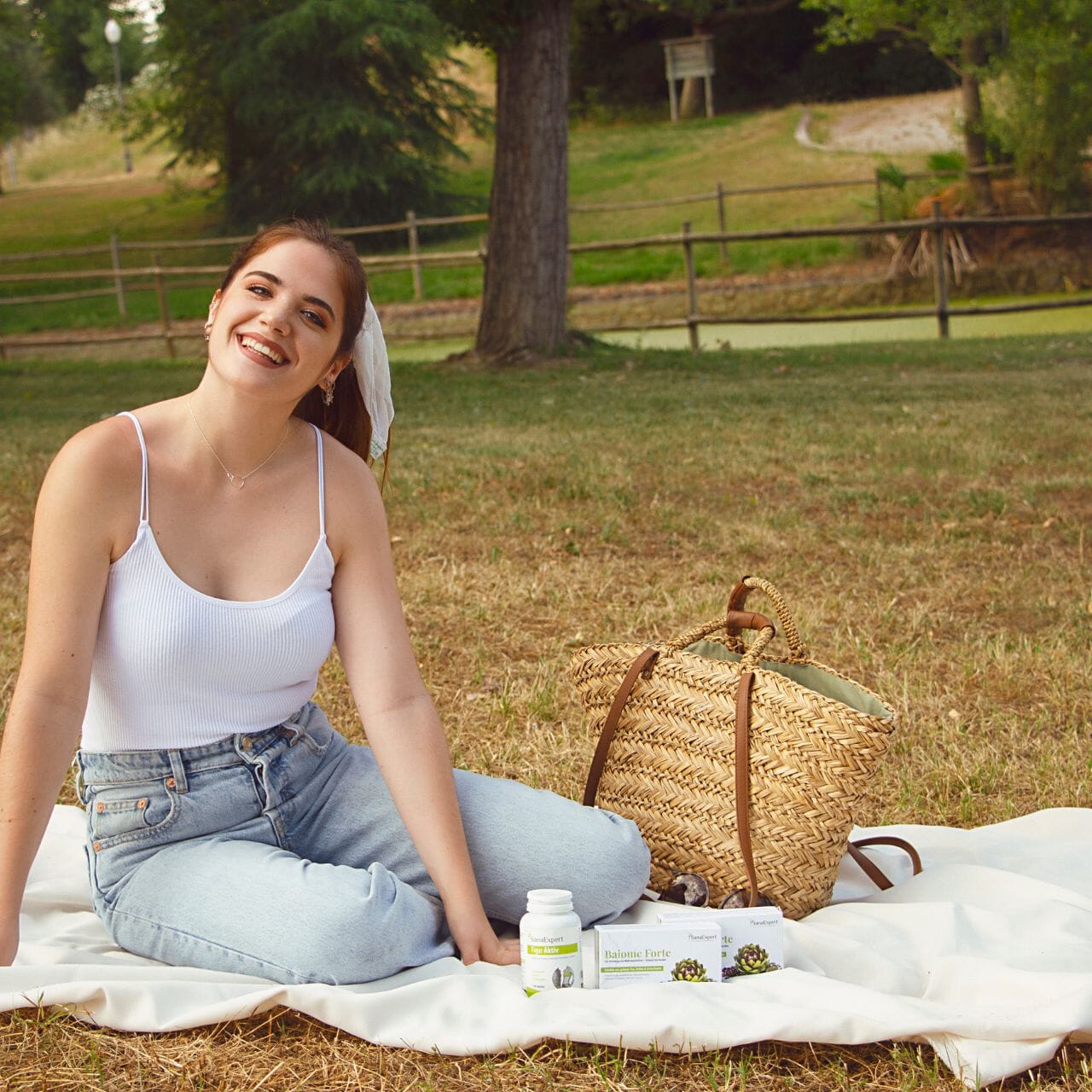 Glückliche junge Frau sitzend auf einem Picknicktuch im Park, lächelnd mit SanaExpert Figur Aktiv und Baïome Forte, entspannte Outdoor-Atmosphäre.
